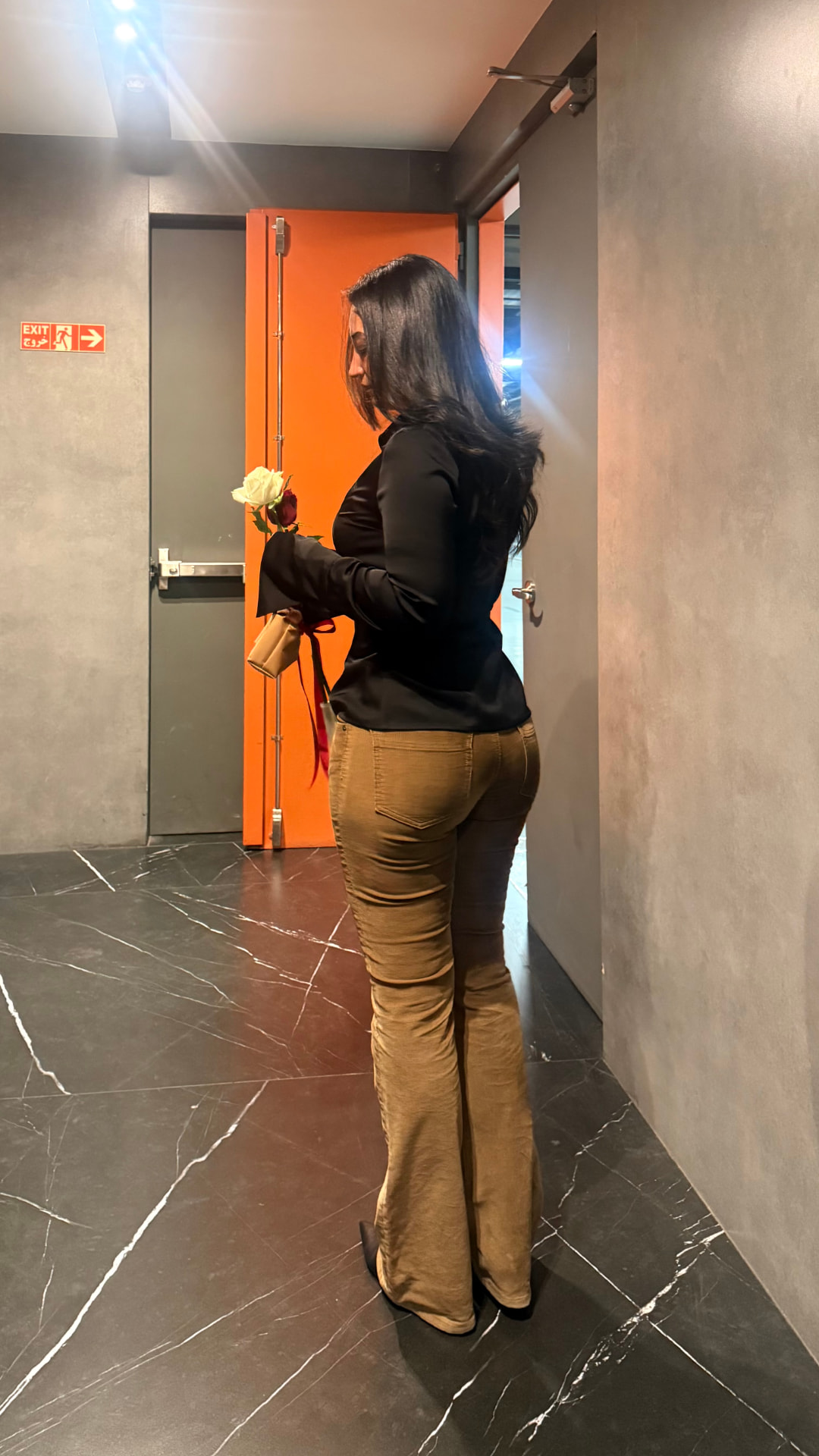 Model in a black satin blouse and camel flared trousers carrying a bouquet through a hallway